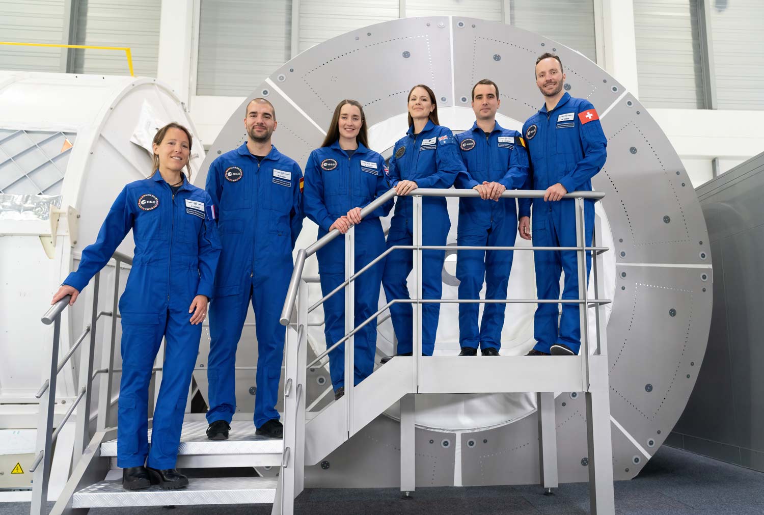 Katherine Bennell-Pegg standing on a staircase with her European Space Agency colleagues in blue jumpsuits and standing in front of space training equipment.